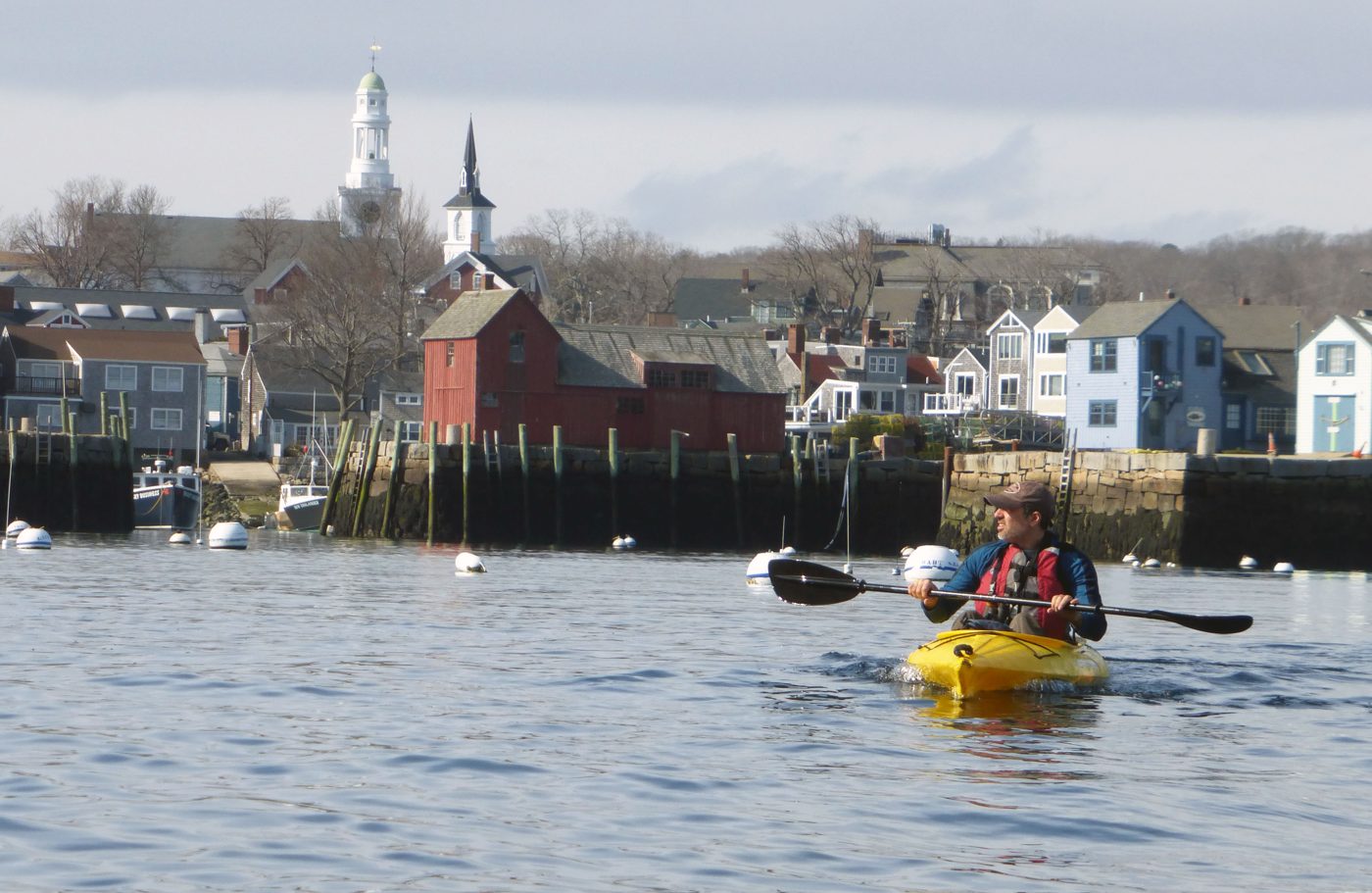 A paddler in a yellow kayak on Rockport Harbor, with red fishing sheds and white steeples behind