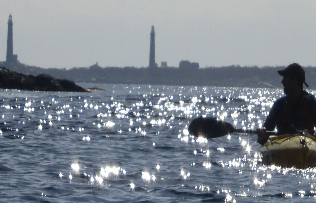 Two granite lighthouse towers on Thacher Island rise over sparkling water, with a kayaker at the right edge