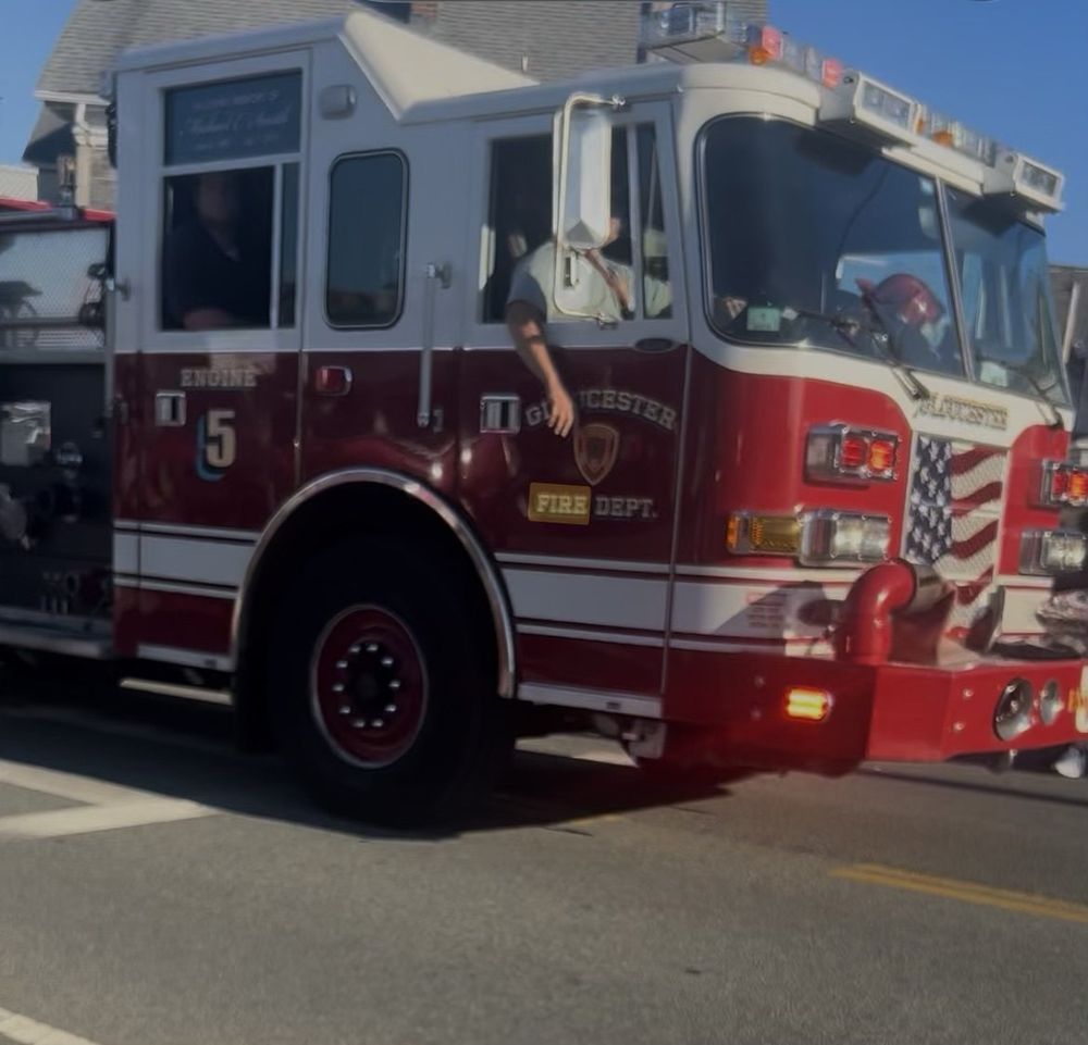 Gloucester Fire Department Engine 5, a red and white fire engine with an American flag painted on its front grille, rolling through an intersection