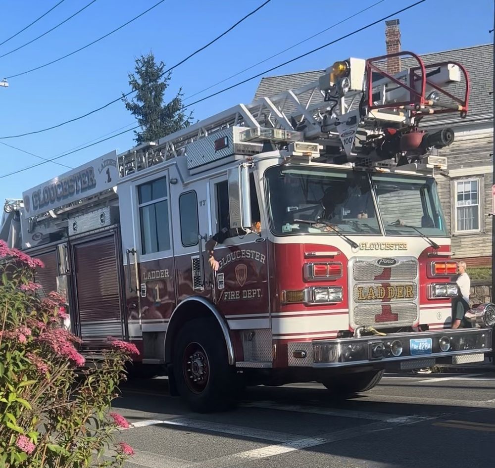Gloucester Fire Department Ladder 1, a red and white Pierce ladder truck with a 100 foot aerial ladder, driving down a sunny Gloucester street past pink flowers