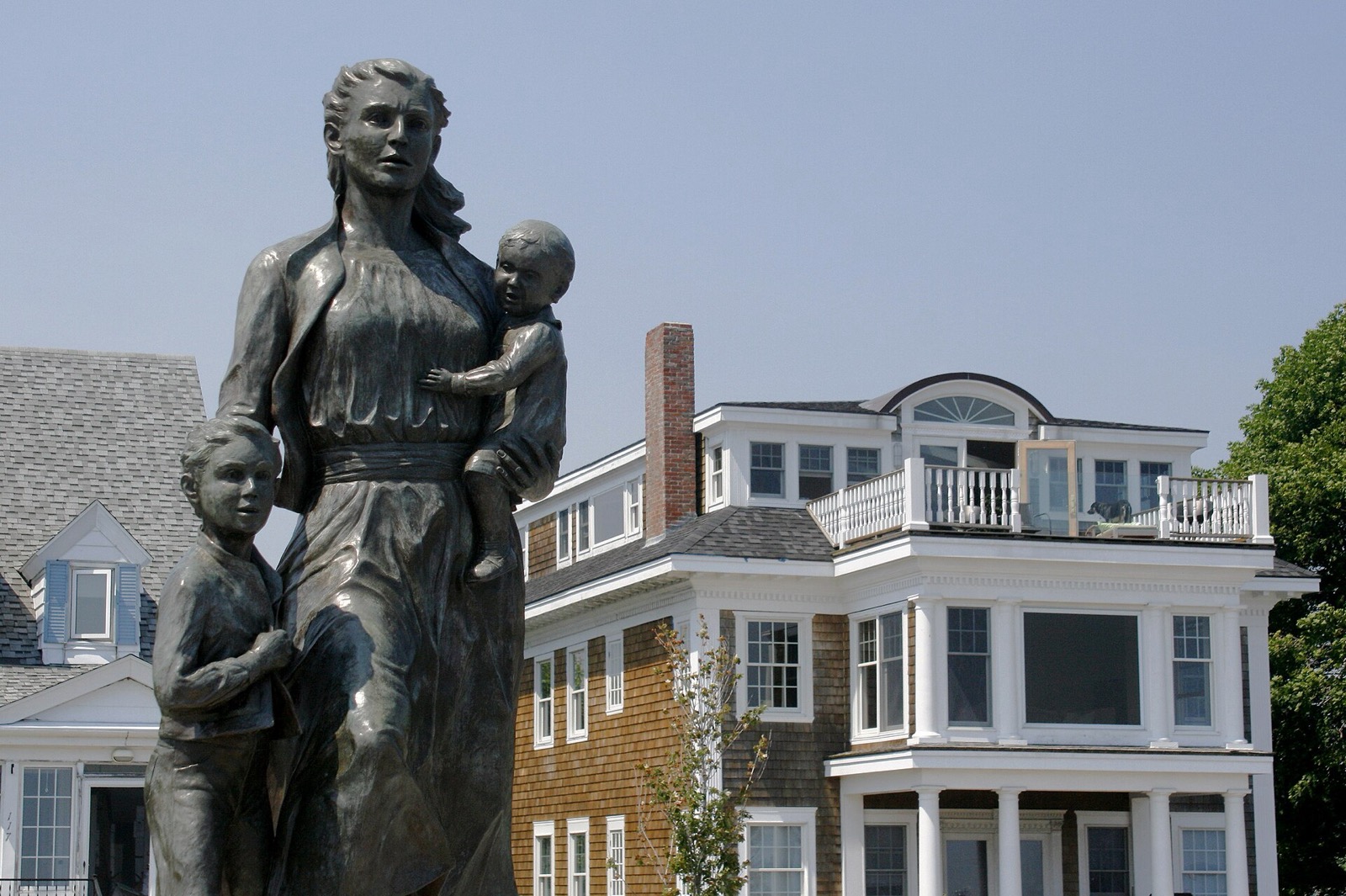 Bronze statue of a woman holding a small child, with an older child at her side, looking out to sea in front of shingled houses on Stacy Boulevard