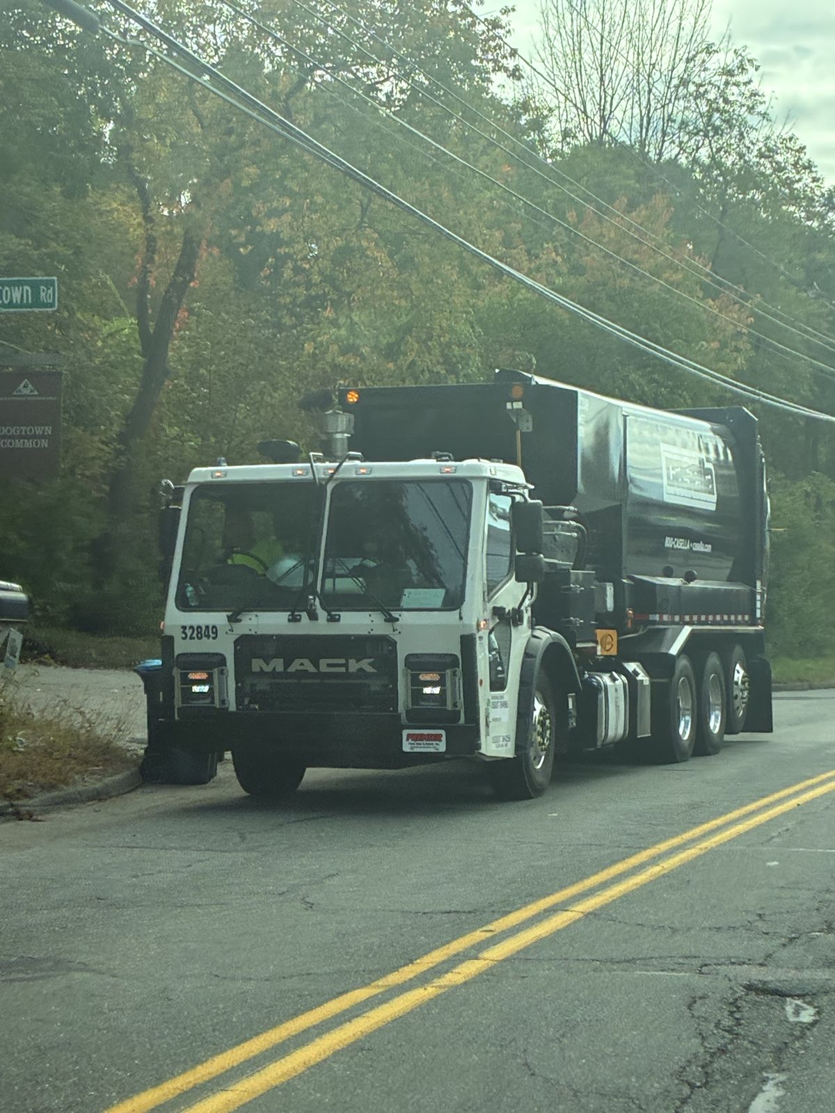 A white and black Casella automated trash truck stops at the curb beside a Dogtown Common sign, with a recycling cart at the roadside
