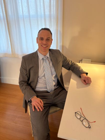 Gil Frieden, in a gray suit and tie, sits at a white desk and smiles