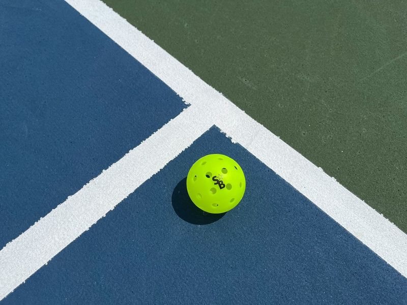 A bright yellow pickleball on a blue and green outdoor court with white lines
