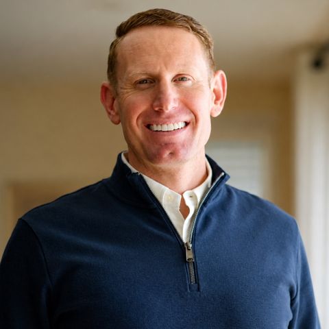 Micah Quinney Jones, in a navy quarter zip sweater over a white collared shirt, smiles at the camera