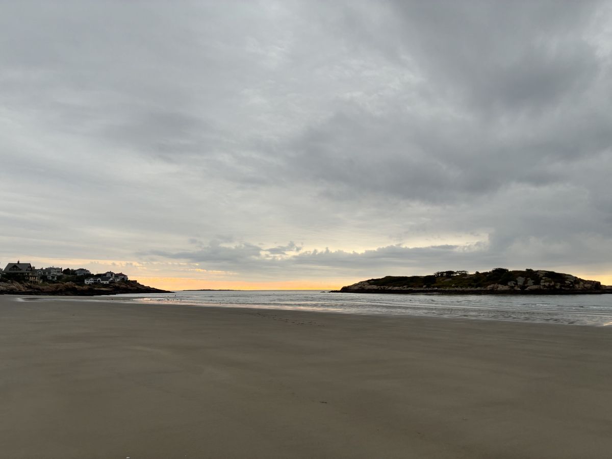 Wide, empty sand at Good Harbor Beach at low tide under heavy gray clouds, with Salt Island on the right and houses on the rocks at the left edge
