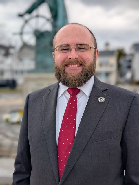 Dru Tarr, in a gray suit and red tie, smiles outdoors on the Gloucester waterfront