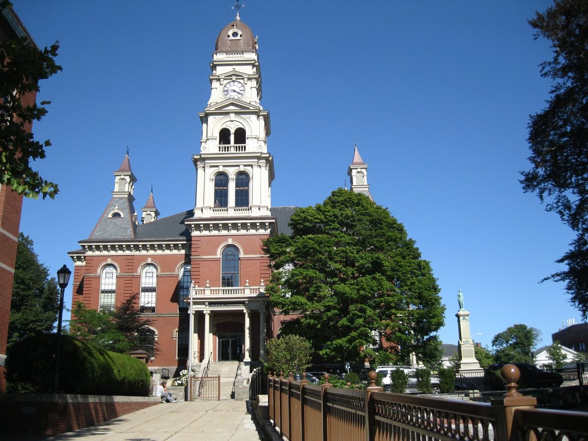 Gloucester City Hall, un edificio de ladrillo rojo con una torre de reloj blanca y alta, bajo un cielo azul despejado, con árboles y un monumento a los caídos a un lado