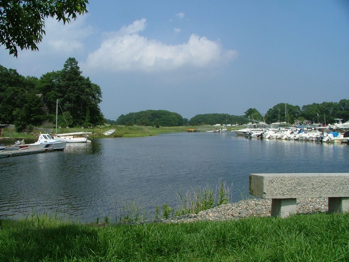Calm blue water on the Essex River in Essex, with small boats at docks on the right, green marsh and trees on the far bank and a stone bench on the grass in front