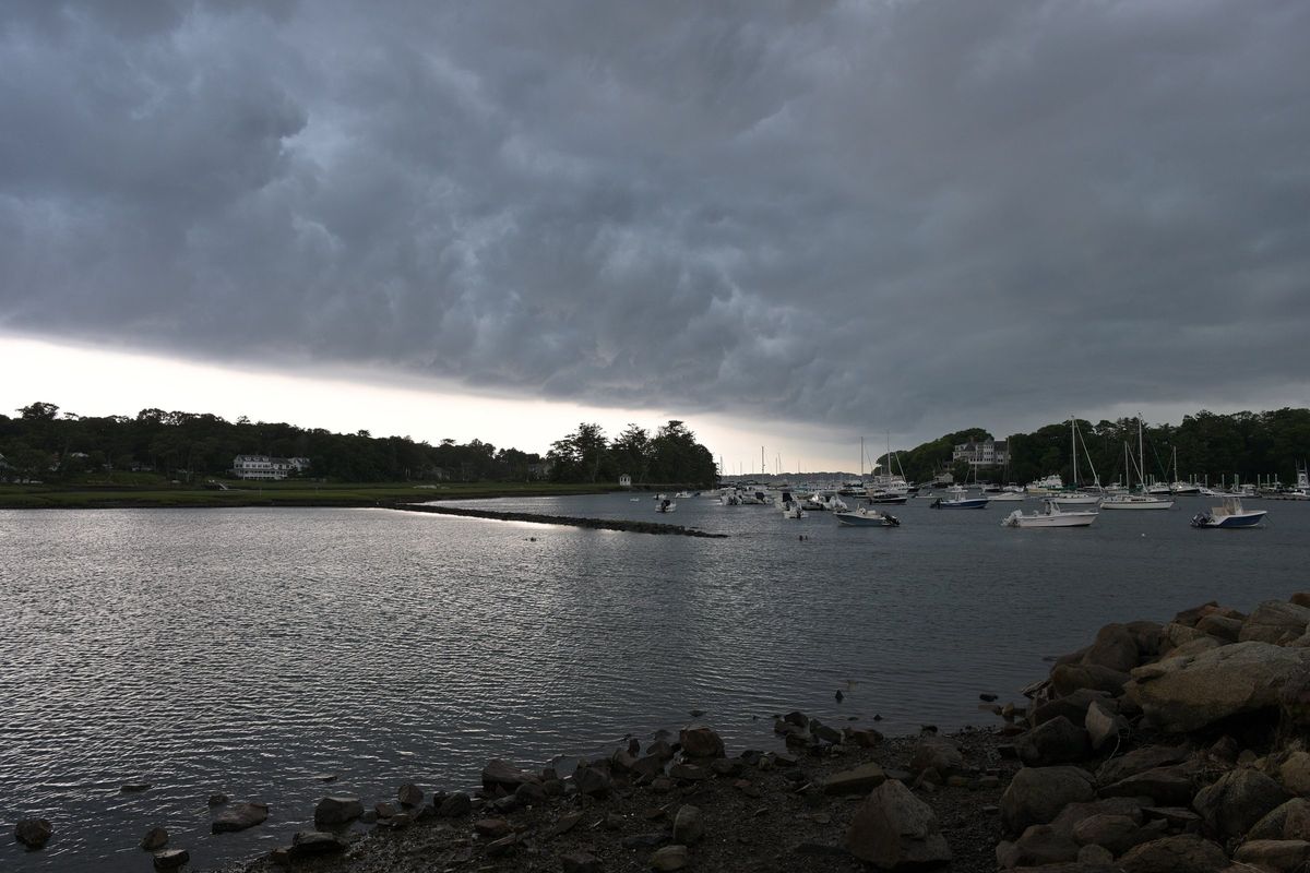 Dark storm clouds over Manchester Harbor, with small boats on their moorings, a stone jetty in calm gray water and rocks along the shore in front